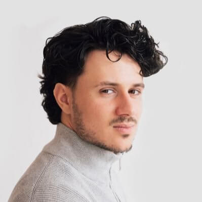 A person with curly black hair and a short beard, wearing a gray high-collar sweater, looks towards the camera against a plain white background, embodying the creativity you'd expect from Toronto's top content creation agency. Conte Studios