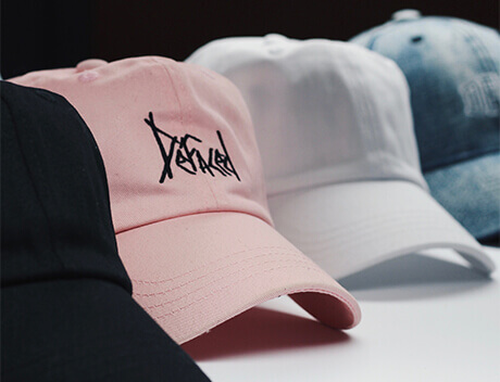 Close-up of four baseball caps in black, pink, white, and blue. The pink cap features "Defined" in stylized black text. Designed by Conte Studios, these caps embody the vibrant creativity of a Toronto-based content creation agency. Conte Studios