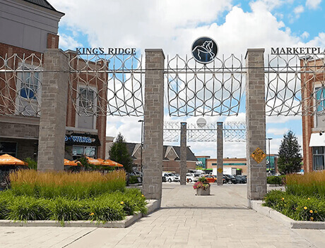 Entrance to King's Ridge Marketplace, designed by Conte Studios, features stone pillars and metal signage. Amidst the surrounding buildings and greenery, the scene unfolds under a blue sky dotted with clouds. Conte Studios