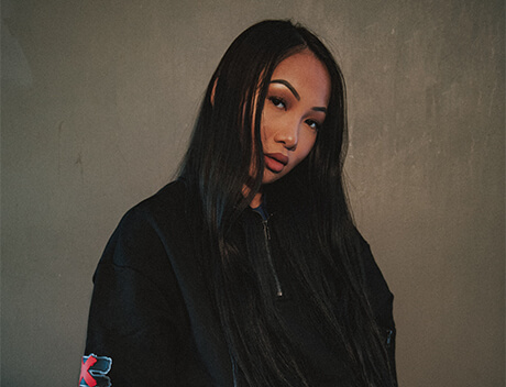 A person with long black hair in a black jacket stands against a gray backdrop, gazing at the camera, embodying the bold aesthetic of a Toronto Design Agency. Conte Studios