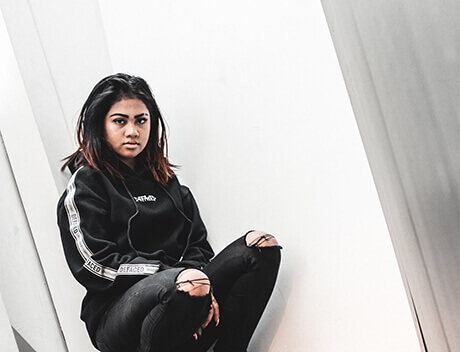 A person with long dark hair in a black sweatshirt and ripped jeans sits against a white angled wall, looking directly at the camera, embodying the edgy creativity of a Toronto Design Agency. Conte Studios