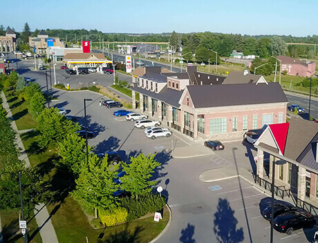 Aerial view of a suburban shopping area with parking spaces, retail buildings, and a few cars. Green trees line the sidewalks, and a fast-food restaurant is visible in the distance. Nearby, a brand design agency from Toronto offers creative flair to local businesses. Conte Studios