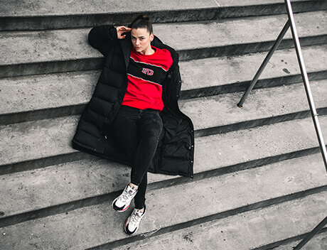 A person in a red shirt and black coat lies on the concrete steps, holding their head with one hand, just outside Conte Studios. Conte Studios