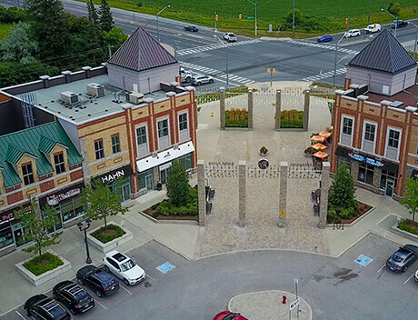 Aerial view of a shopping center, featuring multiple storefronts and parked cars around a central courtyard. Nearby, the intersection buzzes with activity against a backdrop of green fields. Imagine the vibrant scene captured by a Toronto design agency for their content creation showcase. Conte Studios