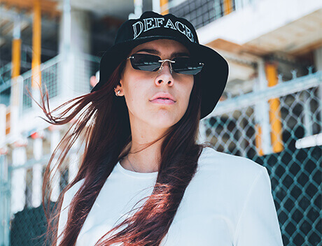 A person wearing sunglasses and a black hat with "DEFACE" text stands in front of a construction site, exuding the edgy vibe of a Toronto Design Agency's latest campaign. The chain-link fence adds an industrial touch to this urban scene. Conte Studios