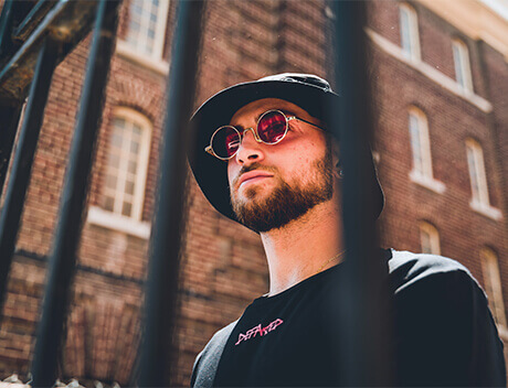 A person wearing a black hat and round sunglasses stands behind a metal fence in front of a brick building, reminiscent of the creative energy at a Toronto Design Agency. Conte Studios