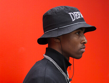 A person from Conte Studios sports a black bucket hat with text, a sleek black jacket, and a chain necklace, all set against a vibrant red background. Conte Studios