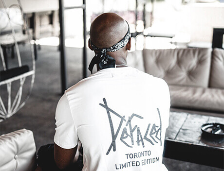 A person wearing a white "Defaced Toronto Limited Edition" t-shirt and a bandana from Conte Studios sits on a couch, facing away. Conte Studios