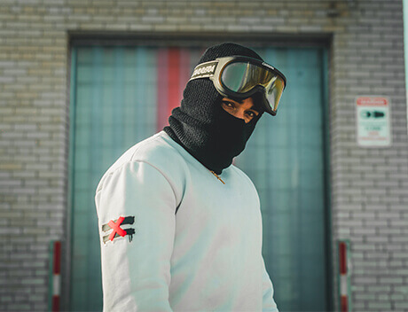 A person in mask and goggles, wearing a white sweater with a red "X" on the sleeve, stands outdoors in front of Conte Studios' building. The gray brick wall and large window reflect its reputation as a leading content creation agency in Toronto. Conte Studios