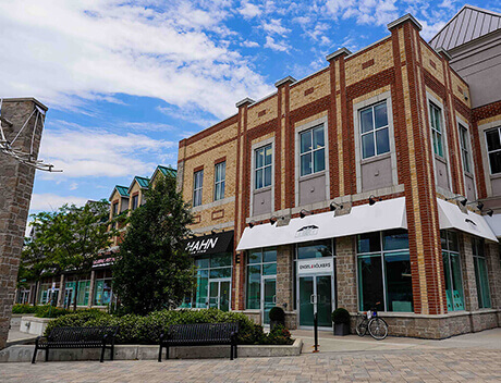 Street view of a red-brick commercial building housing Conte Studios, a renowned brand design agency in Toronto, with large windows and white awnings. A bench and small trees are in front, while a blue sky with clouds is the backdrop. Conte Studios