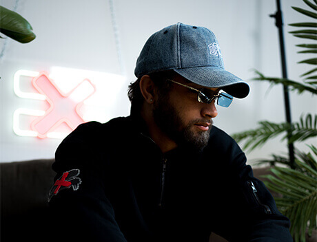 A man wearing a cap and sunglasses sits indoors near a neon X sign with lush plants in the background, embodying the creative vibe of a Toronto Design Agency. Conte Studios