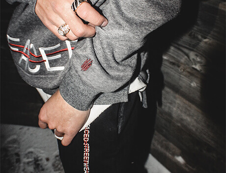 A person stands outdoors near a wooden surface, donning a Conte Studios gray hoodie with red and white text. Black pants with matching side details complement several rings on their fingers, showcasing Toronto's urban flair. Conte Studios