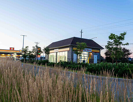 A small car wash building, crafted with the finesse of a Toronto Design Agency, is nestled among plants and trees. In the background, a gas station is faintly visible at dusk, echoing the creativity often seen at Conte Studios. Conte Studios