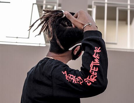 In an indoor setting, a person in a black sweatshirt with red graffiti-style text on the sleeve, possibly from Conte Studios, adjusts their hair while wearing a mask. Conte Studios