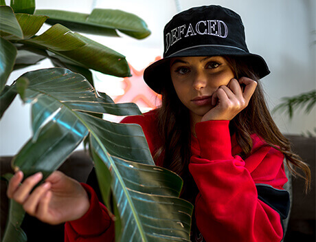 Person in a red sweater and black hat rests their head on their hand, sitting near large green leaves indoors. This serene moment captures the creative spirit often embraced at a Toronto Design Agency, where inspiration thrives amid nature's beauty. Conte Studios