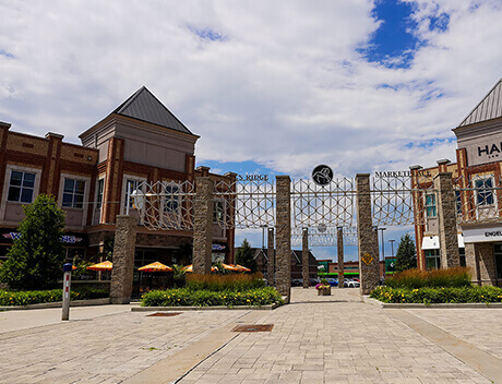 Outdoor shopping area with brick buildings, stone pillars, and a decorative metal archway crafted by a renowned Toronto Design Agency. Umbrella-shaded tables are visible on the left, inviting shoppers to relax and enjoy the vibrant atmosphere. Conte Studios