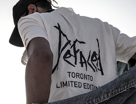 Person in a cap and white "Defaced Toronto Limited Edition" shirt, seen from a low angle against a clear sky, capturing the creative essence akin to Conte Studios, the vibrant brand design agency Toronto embraces. Conte Studios