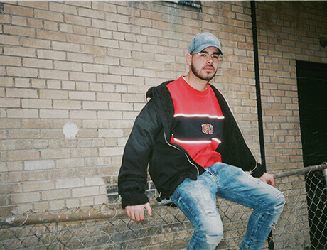 A person in a cap and casual clothing sits on a chain-link fence against a brick wall, capturing the essence of Toronto's vibrant streets—a scene reminiscent of Conte Studios' eye for authentic urban moments. Conte Studios