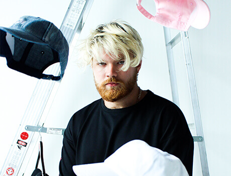 A man with blond hair and a beard stands in front of a ladder, wearing a black shirt. Several caps are suspended around him, showcasing the creative flair of Conte Studios, a renowned brand design agency in Toronto. Conte Studios