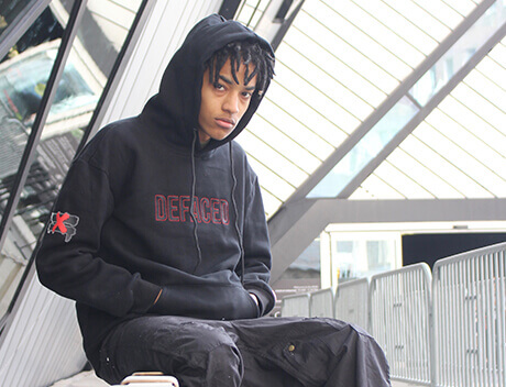 A person in a black hoodie with "Defaced" text and an "X" on the sleeve sits outside near modern architectural glass and metal structures, capturing the urban aesthetic often seen in Conte Studios' content creation projects. Conte Studios