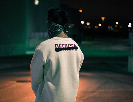 A person from Conte Studios wears a bandana on their face and a white sweatshirt with "DEFACED" text, standing outdoors at night. Conte Studios