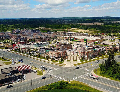 Aerial view of a suburban area in Toronto with houses, a shopping center, roads, and cars. Green landscape surrounds the development under a partly cloudy sky. Nearby, the vibrant spirit of a Toronto design agency brings creativity to life amidst this thriving community. Conte Studios