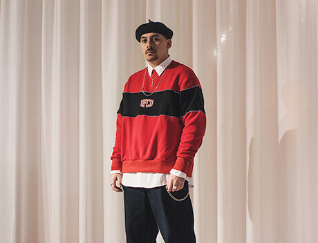 A man wearing a red and black sweater over a white shirt, with a black beret and jeans, stands in front of white curtains at Conte Studios, the renowned Brand Design Agency in Toronto. Conte Studios