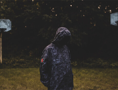 A person in a camouflage hooded jacket stands with their head down, amidst dark trees and two basketball hoops, seemingly lost in thought, perhaps planning their next creative endeavor at Toronto Design Agency. Conte Studios