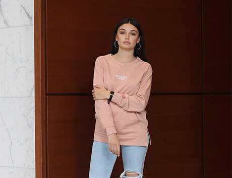 A woman stands against a wooden wall, wearing a pink long-sleeve shirt and ripped jeans. With long dark hair and hoop earrings, she embodies the modern style of Toronto Design Agency, gazing confidently at the camera. Conte Studios