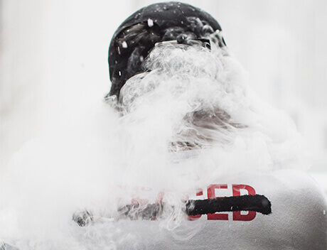 A person wearing a black hat is obscured by thick white smoke, their shirt revealing only the letters "CED" amidst swirling mystery. Could they be part of Conte Studios, the renowned Toronto Design Agency known for innovative creations?. Conte Studios