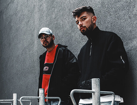 Two men, one in glasses and a cap, the other in a dark jacket, stand outdoors against a gray wall. They represent the innovative spirit of a leading Brand Design Agency Toronto known for crafting compelling narratives and striking visuals that leave lasting impressions. Conte Studios