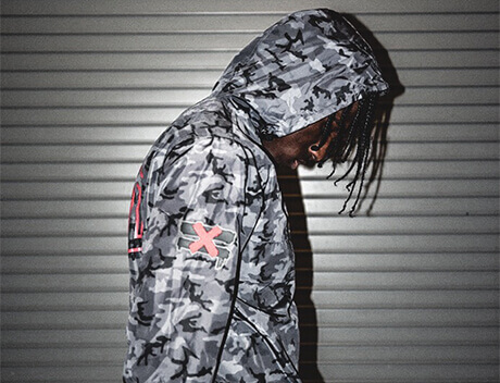 Person wearing a gray camo hooded jacket with an "X" on the sleeve, standing against a corrugated metal background, embodies the urban vibe captured by a top Toronto Design Agency. Conte Studios