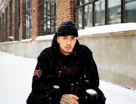 A person in a black hoodie and beanie crouches in a snowy outdoor setting, with a brick building in the background, capturing the essence of winter for Conte Studios, a renowned content creation agency in Toronto. Conte Studios