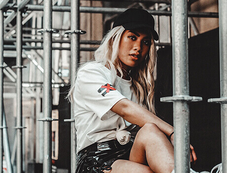 A person with long hair and a black cap sits among metal scaffolding, representing Conte Studios in a white shirt and shiny shorts. Conte Studios