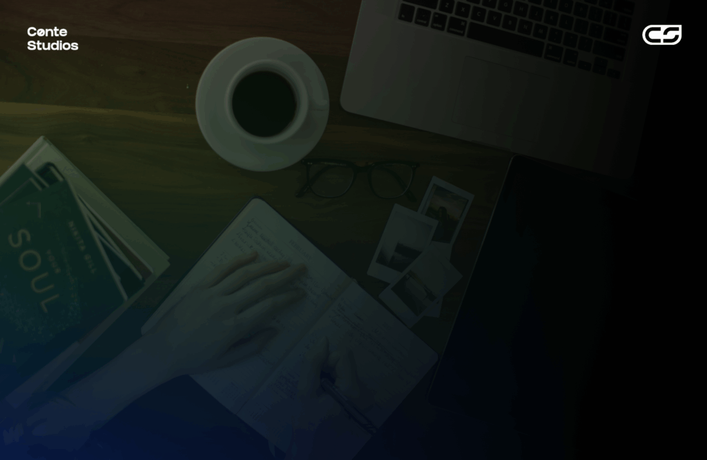 A person writes in a notebook on a wooden desk with a coffee cup, eyeglasses, instant photos, a book, and a laptop nearby. Conte Studios