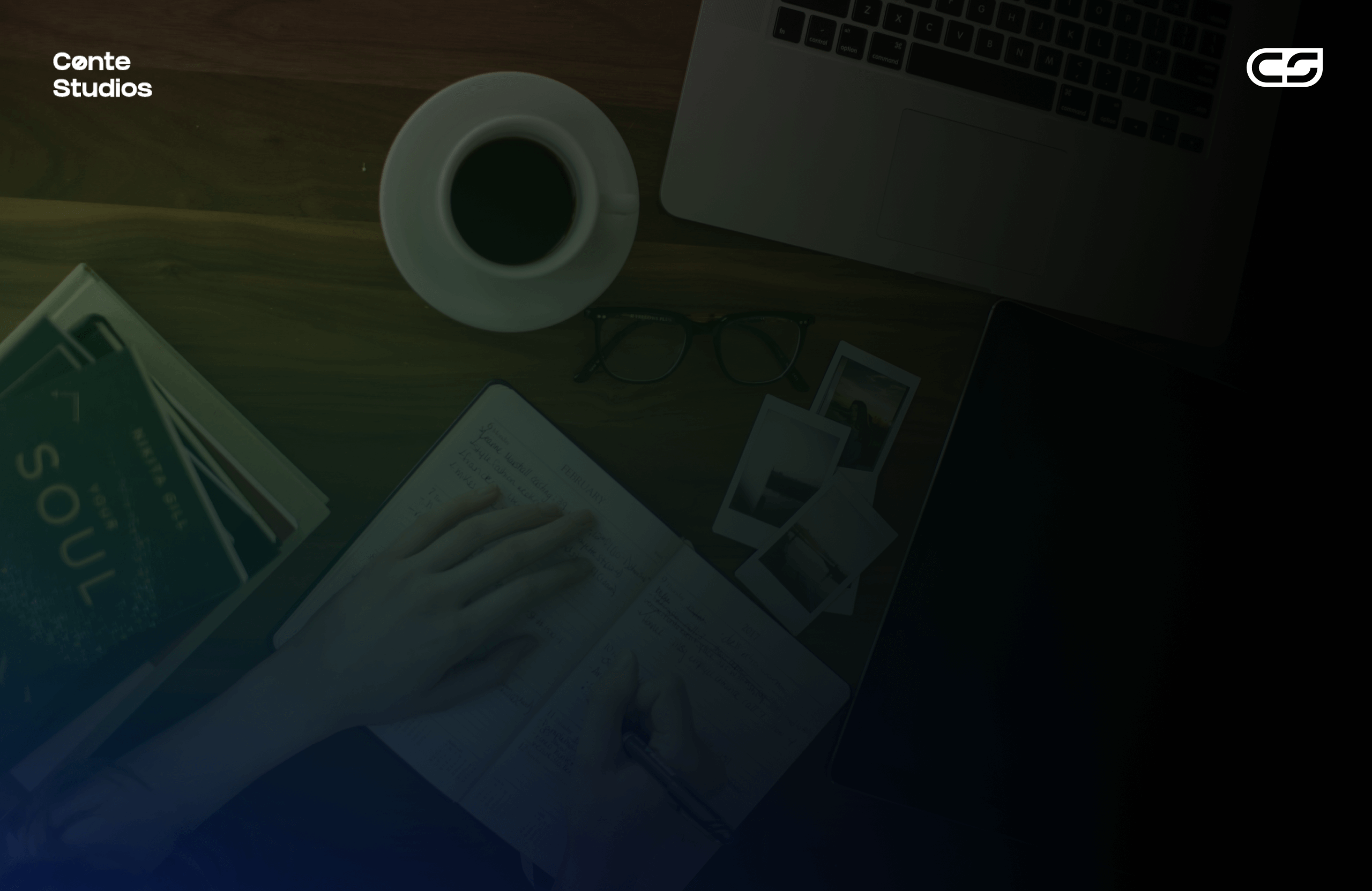A person writes in a notebook on a wooden desk with a coffee cup, eyeglasses, instant photos, a book, and a laptop nearby. Conte Studios