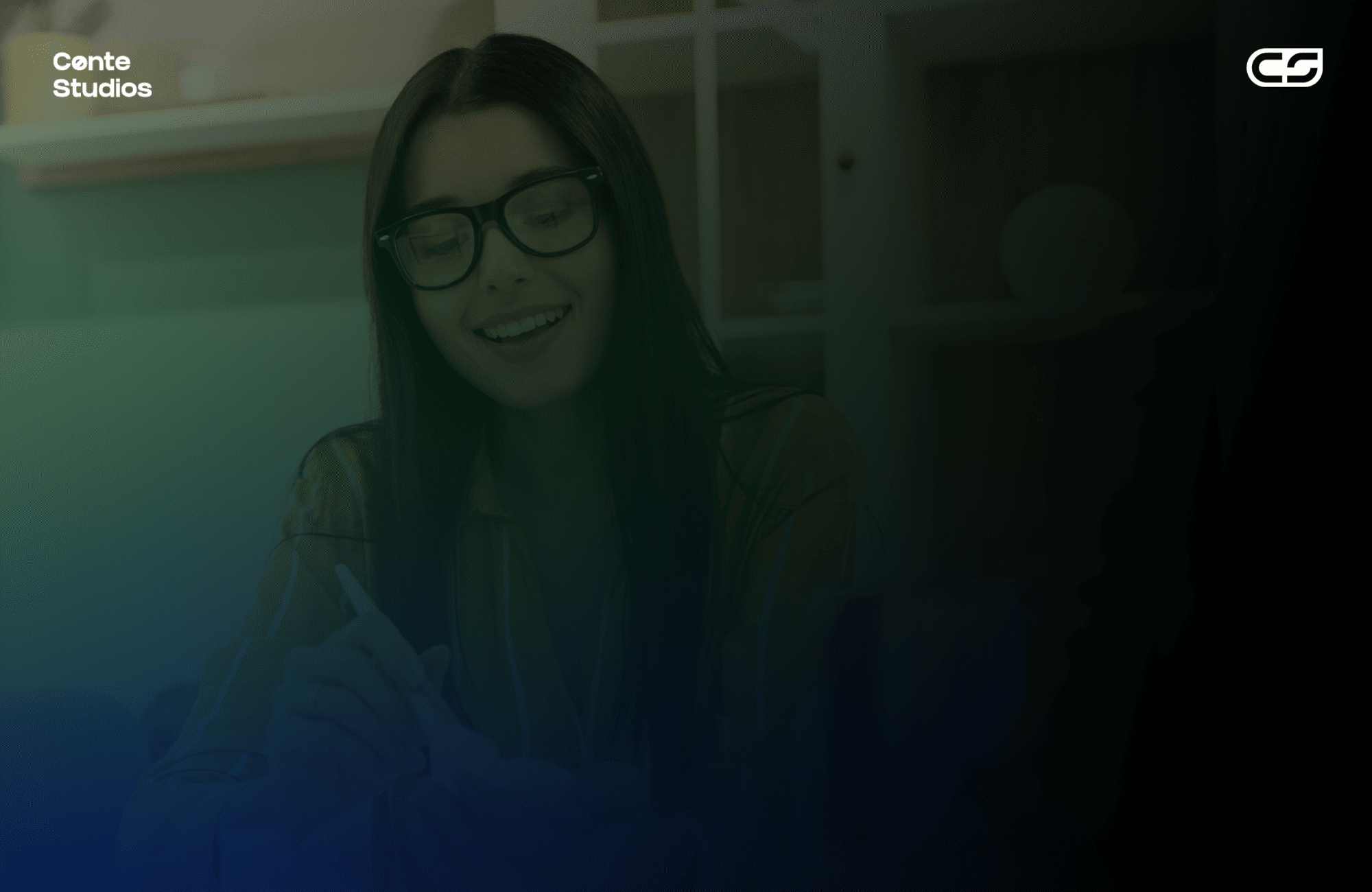 A person with long dark hair and glasses is smiling and using a pen in an office with shelves in the background. Conte Studios