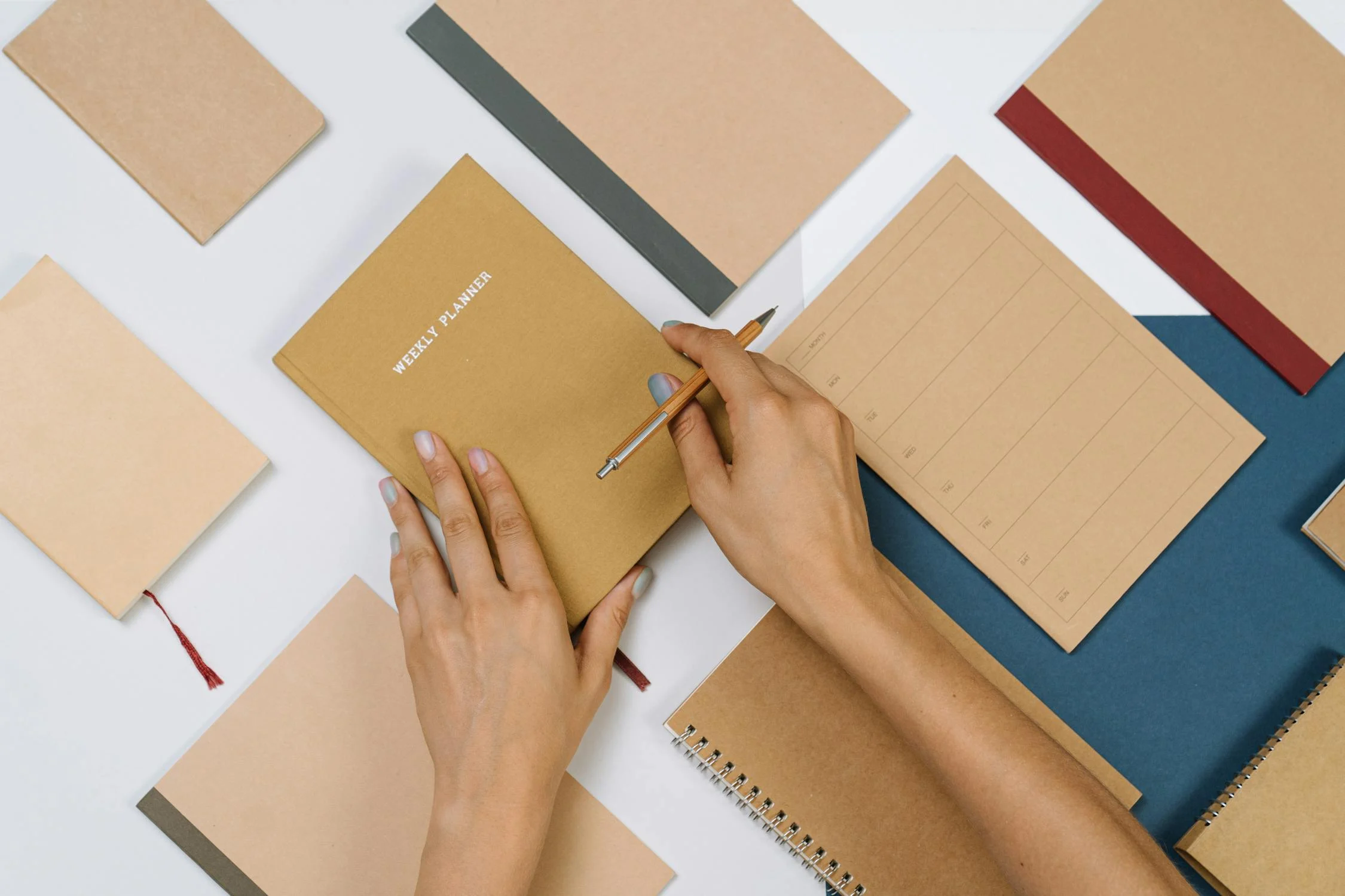 A person holds a brown weekly planner and a pen from Conte Studios, surrounded by various notebooks and notepads on a white surface. Conte Studios
